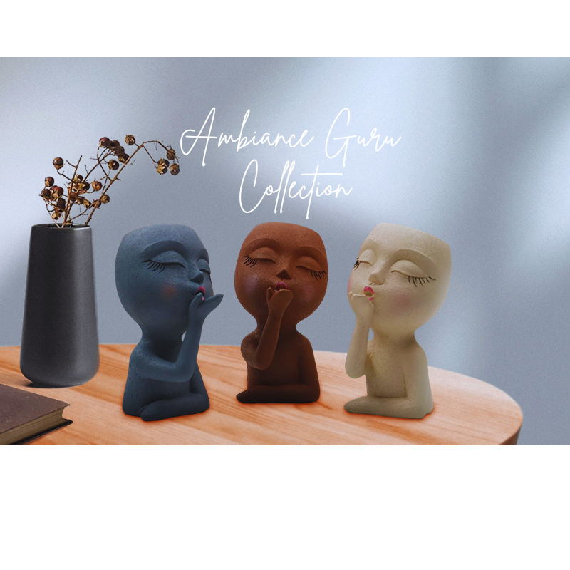 Three ceramic face sculptures on a wooden surface with a vase of dried flowers, labeled 'Ambiance Guru Collection'.